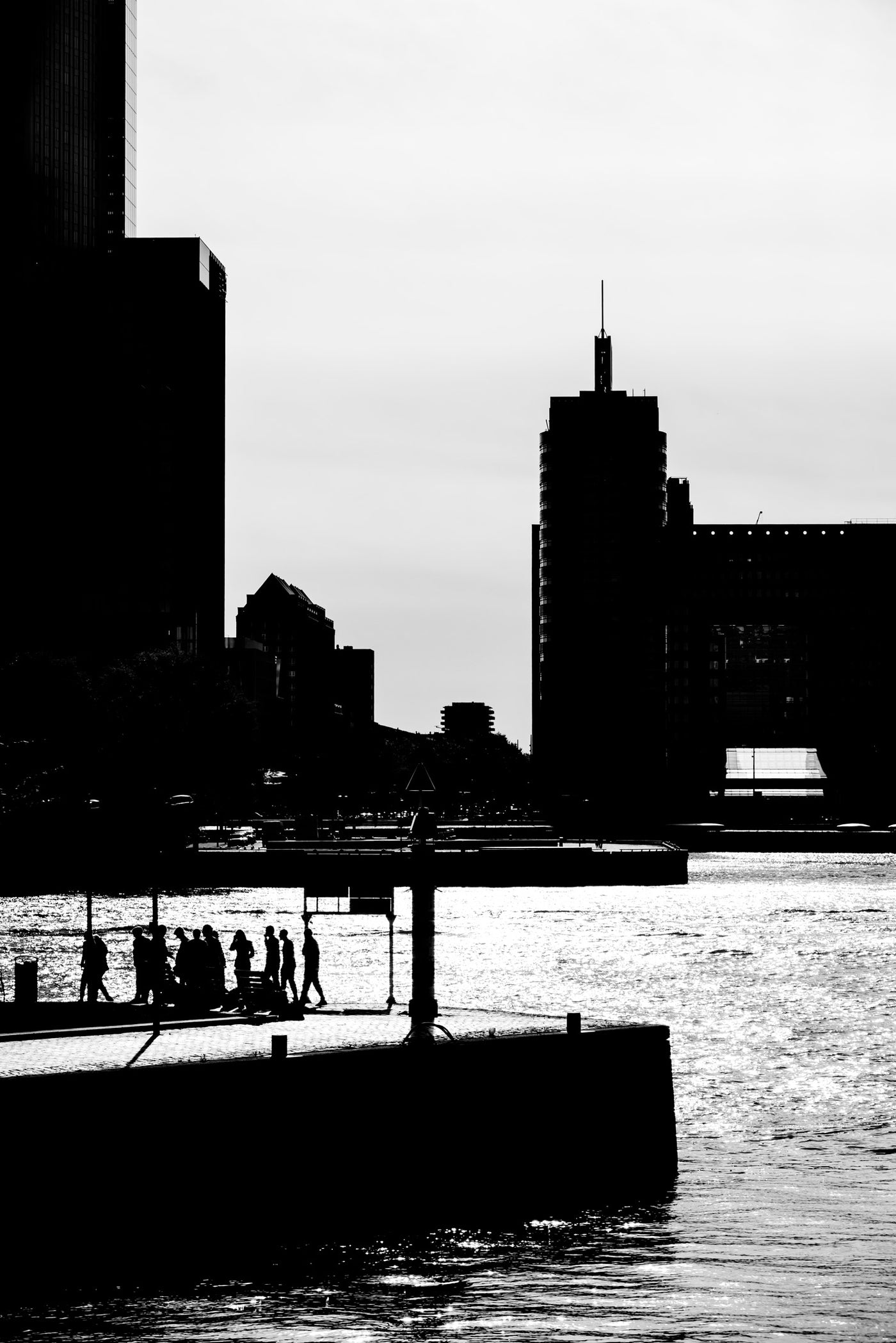 Black and white Rotterdam waterfront photograph with silhouetted buildings, a pier, water, and small walking figures against a bright sky.