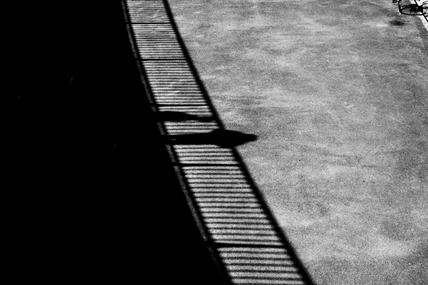 Black and white high-contrast photograph of a long shadow cast across a textured concrete surface at the pathway leading to the Erasmusbrug in Rotterdam