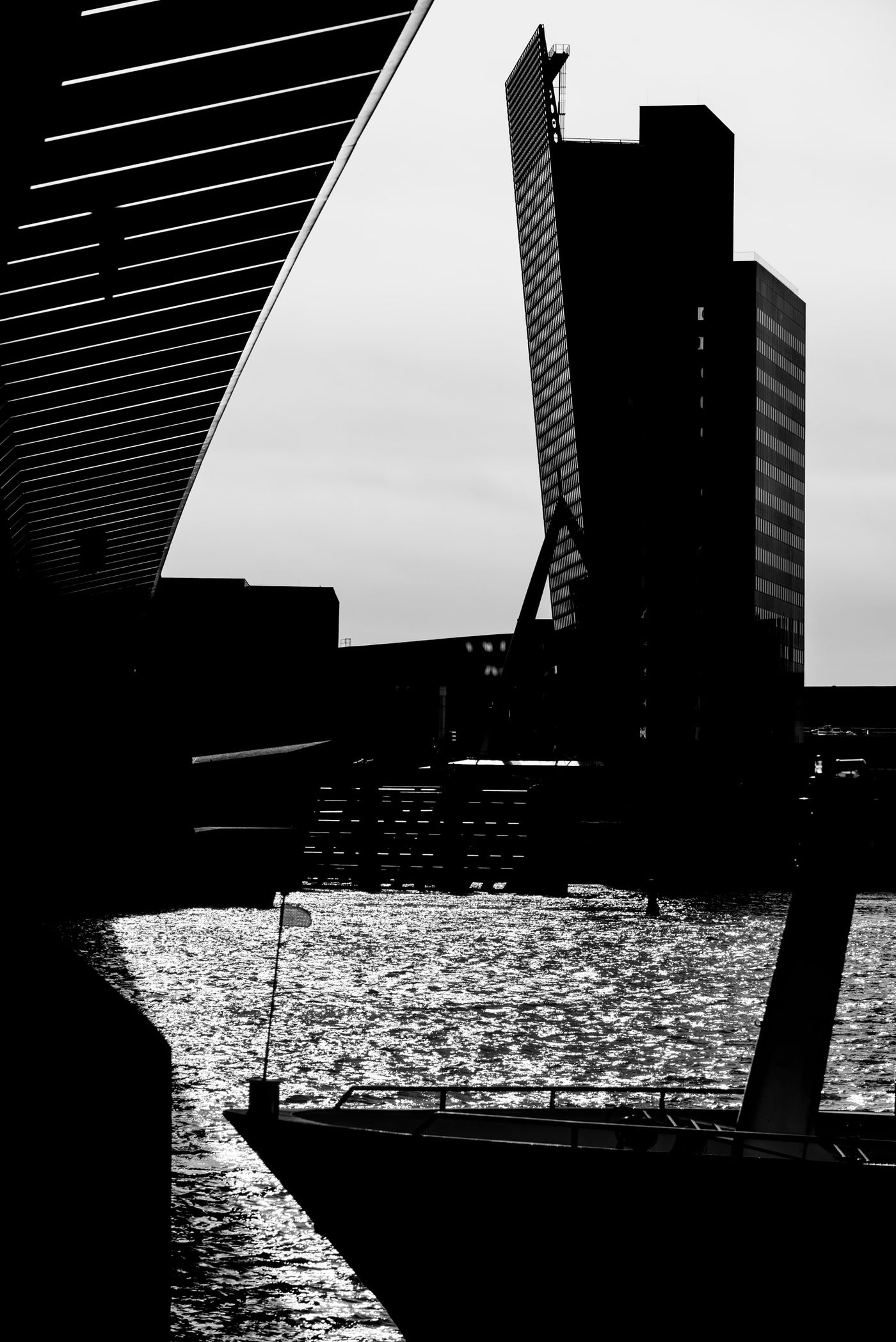 Black and white architectural photograph featuring the Erasmusbrug and surrounding buildings reflected in water of the Maas, with a boat in the foreground.