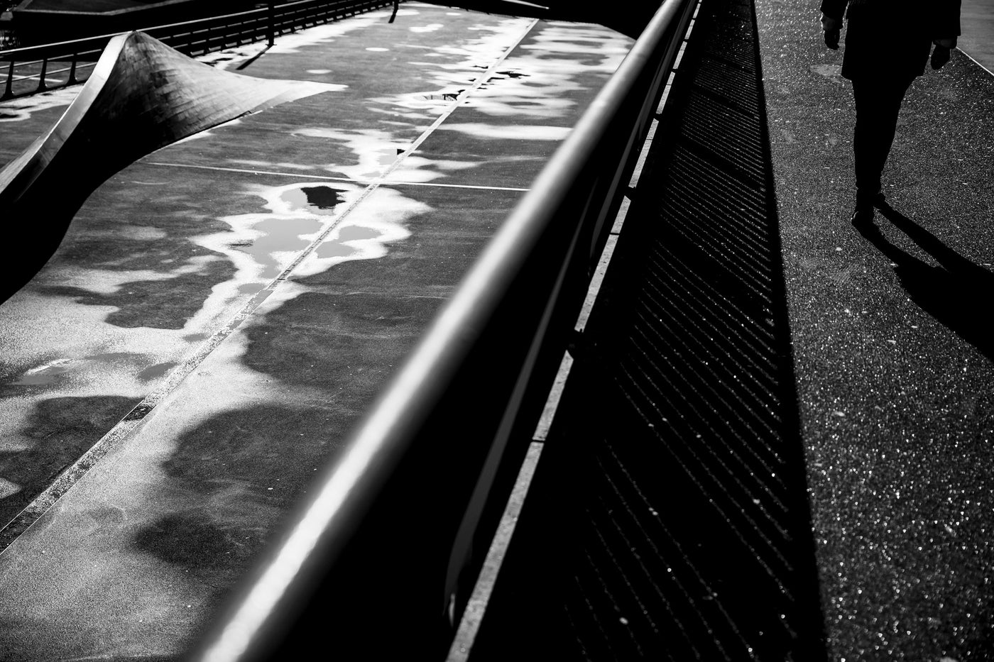 Black and white high-contrast street photograph showing a concrete ramp with wet reflective surfaces, a metal railing, a walkway, and the silhouette of a person walking on the Erasmusbrug in Rotterdam.