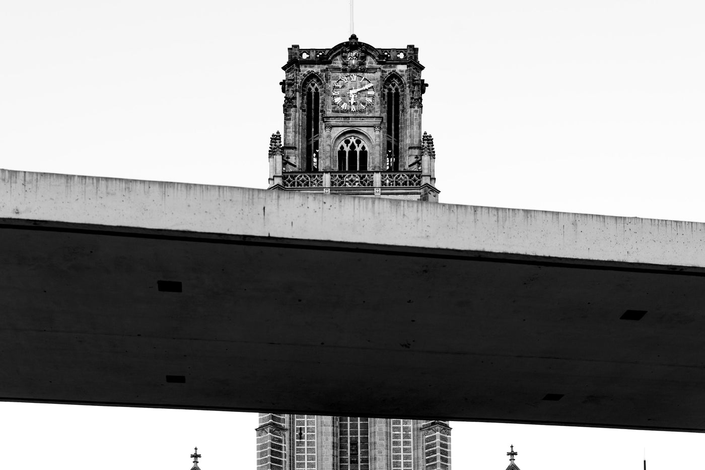 Black and white architectural photograph featuring a clock tower framed by a large concrete bridge in the foreground, with concrete structure visible below in Rotterdam.