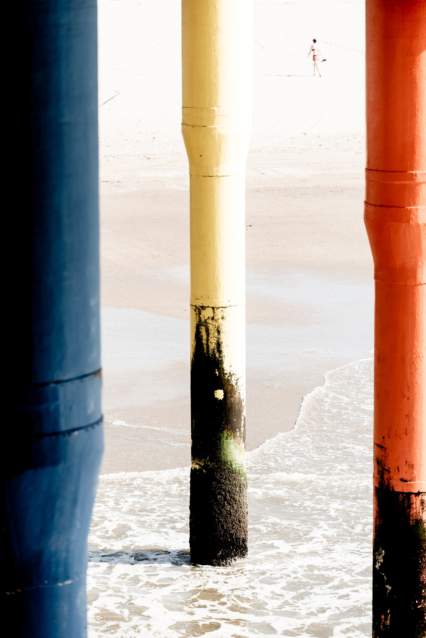 Three large vertical pier columns in blue, cream, and rust orange standing in shallow ocean water at the beach, with pale sand and a distant person visible in the bright background.