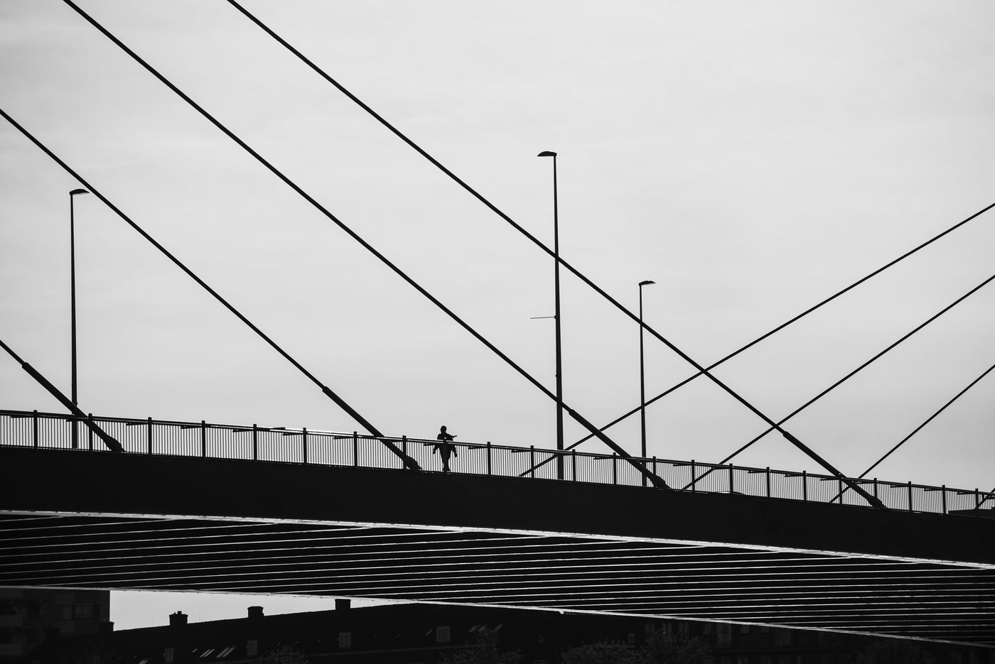 Black and white photograph of a person walking across the Willemsbrug bridge in Rotterdam, framed by the bridge cables and streetlights against a cloudy sky.
