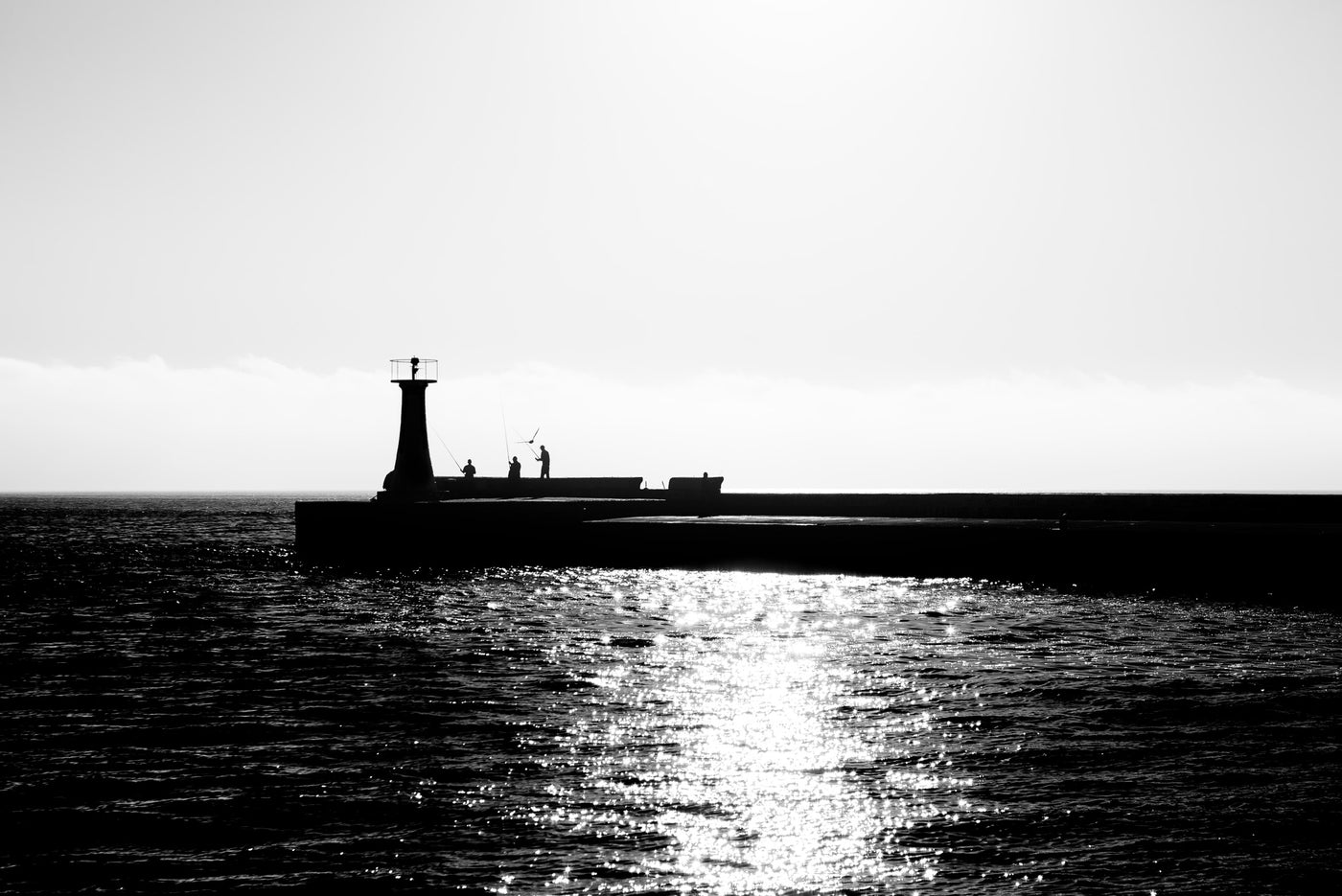 Black and white photograph of a Kalk Bay lighthouse silhouette on a long pier extending into calm water, with bright sunlight reflecting across the surface.