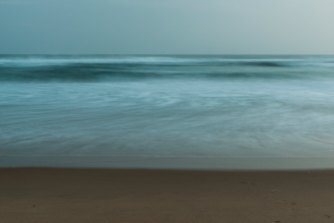 A calm beach scene with gentle blue-gray ocean waves washing onto sandy shore under a pale sky.