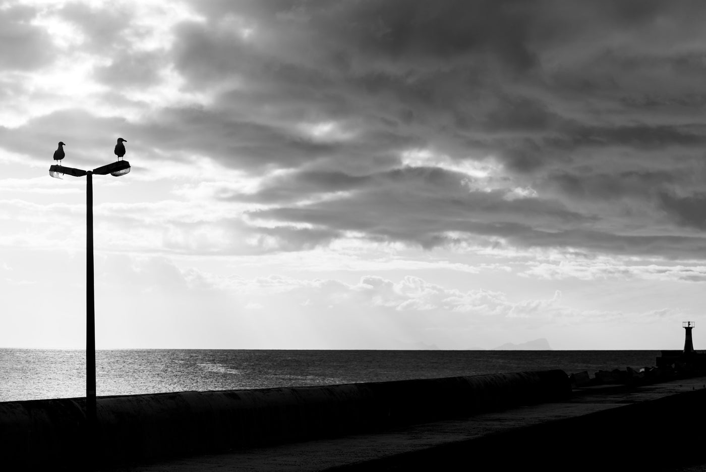 Black and white photograph of a seaside promenade at Kalk bay, Cape Town, with a tall lamp post carrying two birds perched on its top, a long pier extending into calm water, dramatic cloudy sky, and a lighthouse visible in the distance.