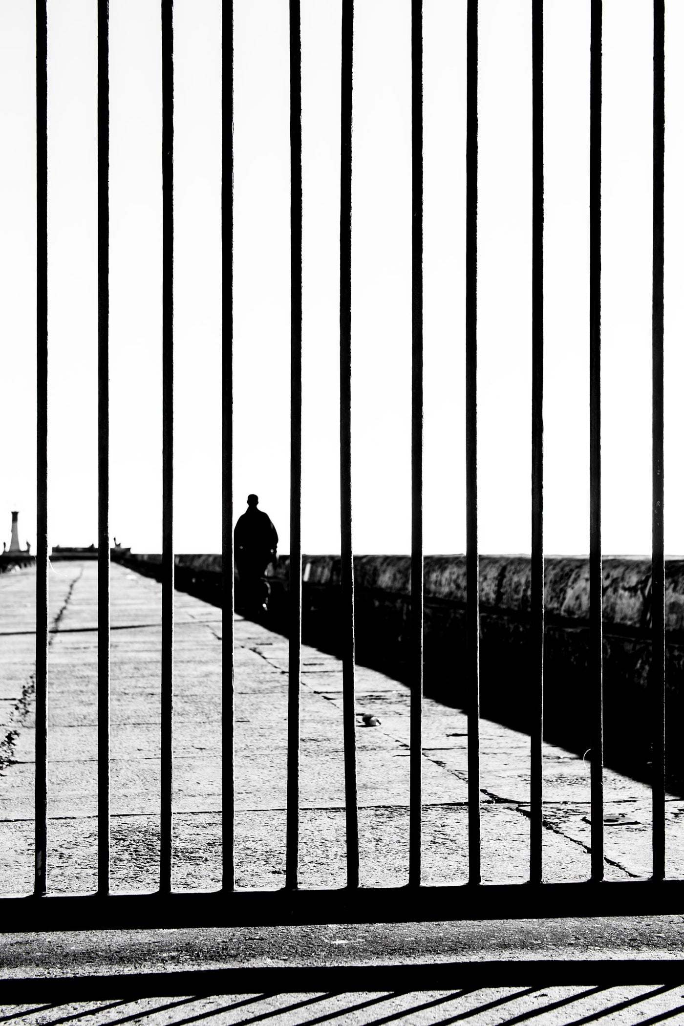 Black and white fine art photograph featuring a silhouetted figure walking along a Kalk Bay pier, framed by a vertical metal gate in the foreground.