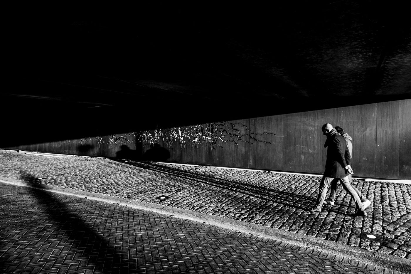 Black and white street photograph showing a person walking along a cobblestone path beside a concrete wall under a dark bridge or overpass.