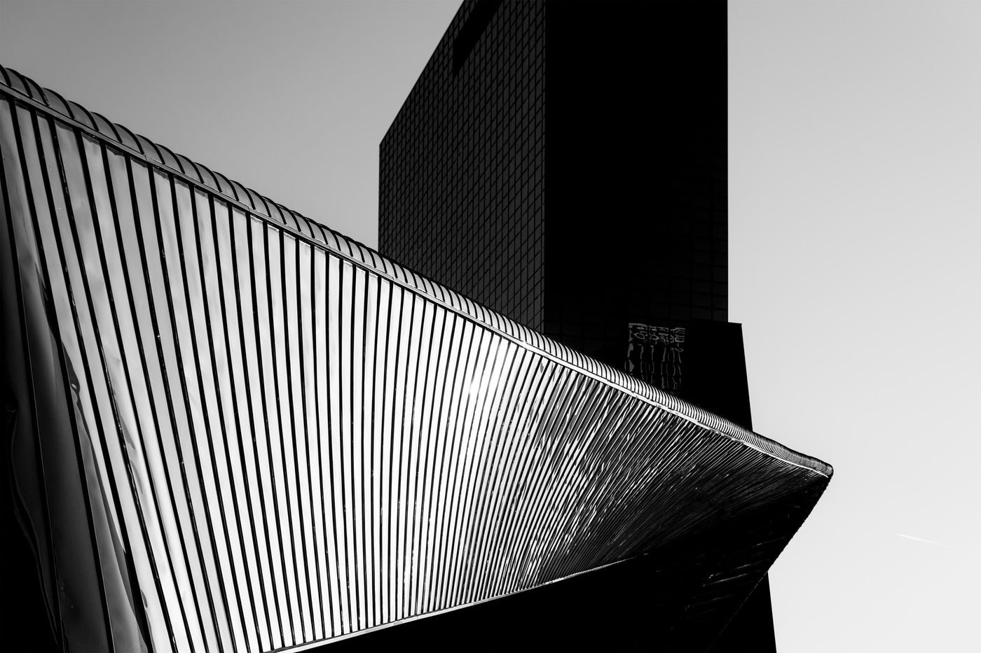 Black and white architectural photograph of modern building facades, featuring a slatted metal roof structure in the foreground and a dark glass skyscraper behind it against a pale sky in central Rotterdam.