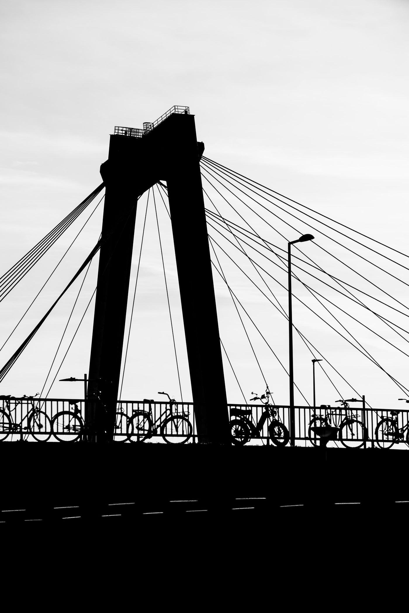 Black and white photograph of a cable-stayed bridge silhouette with bicycles parked along the railing against a pale sky.