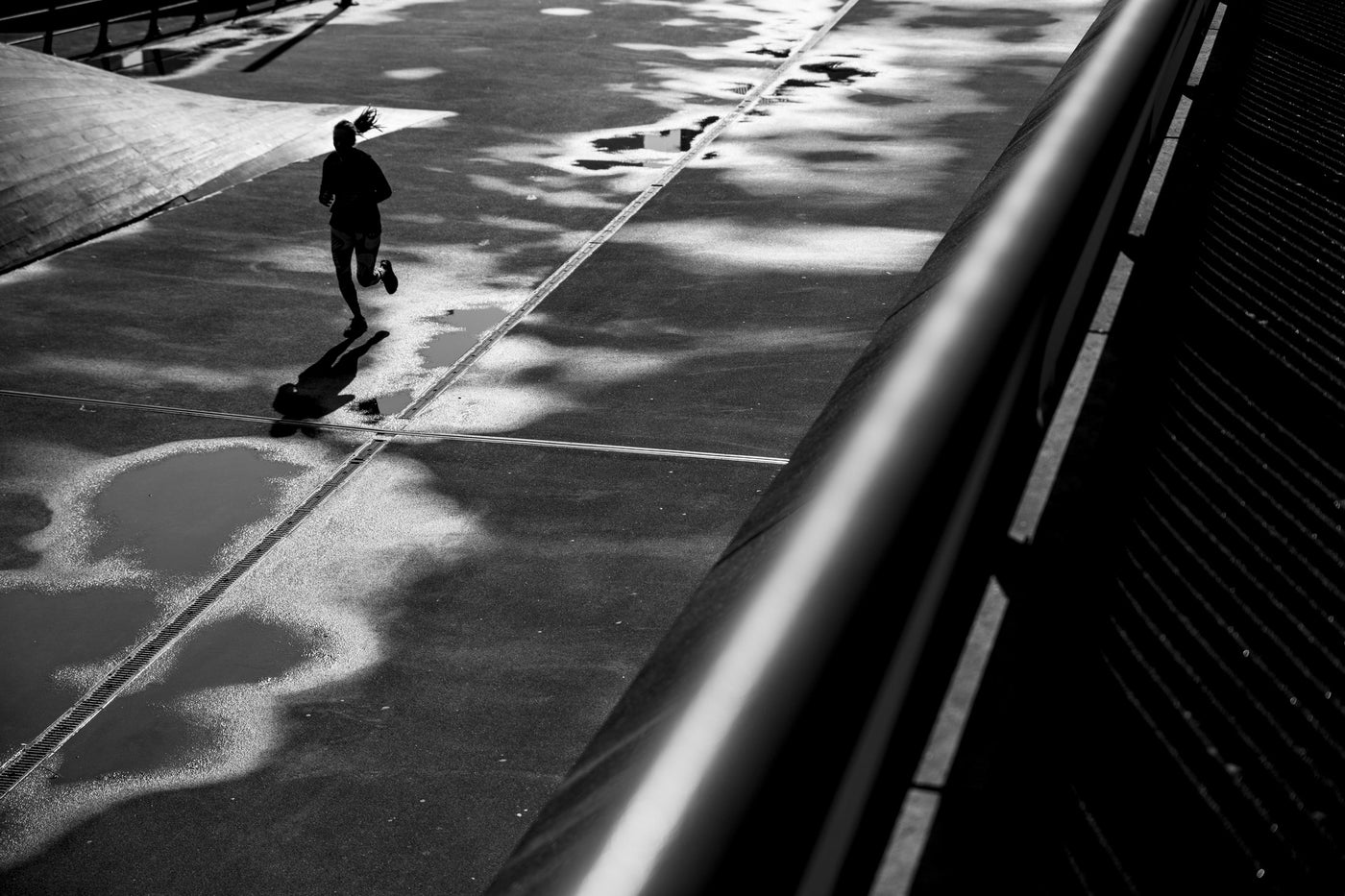 Black and white high-contrast street photograph shot in Rotterdam showing a silhouetted person running across a paved surface with dramatic tree shadows and a dark railing in the foreground.