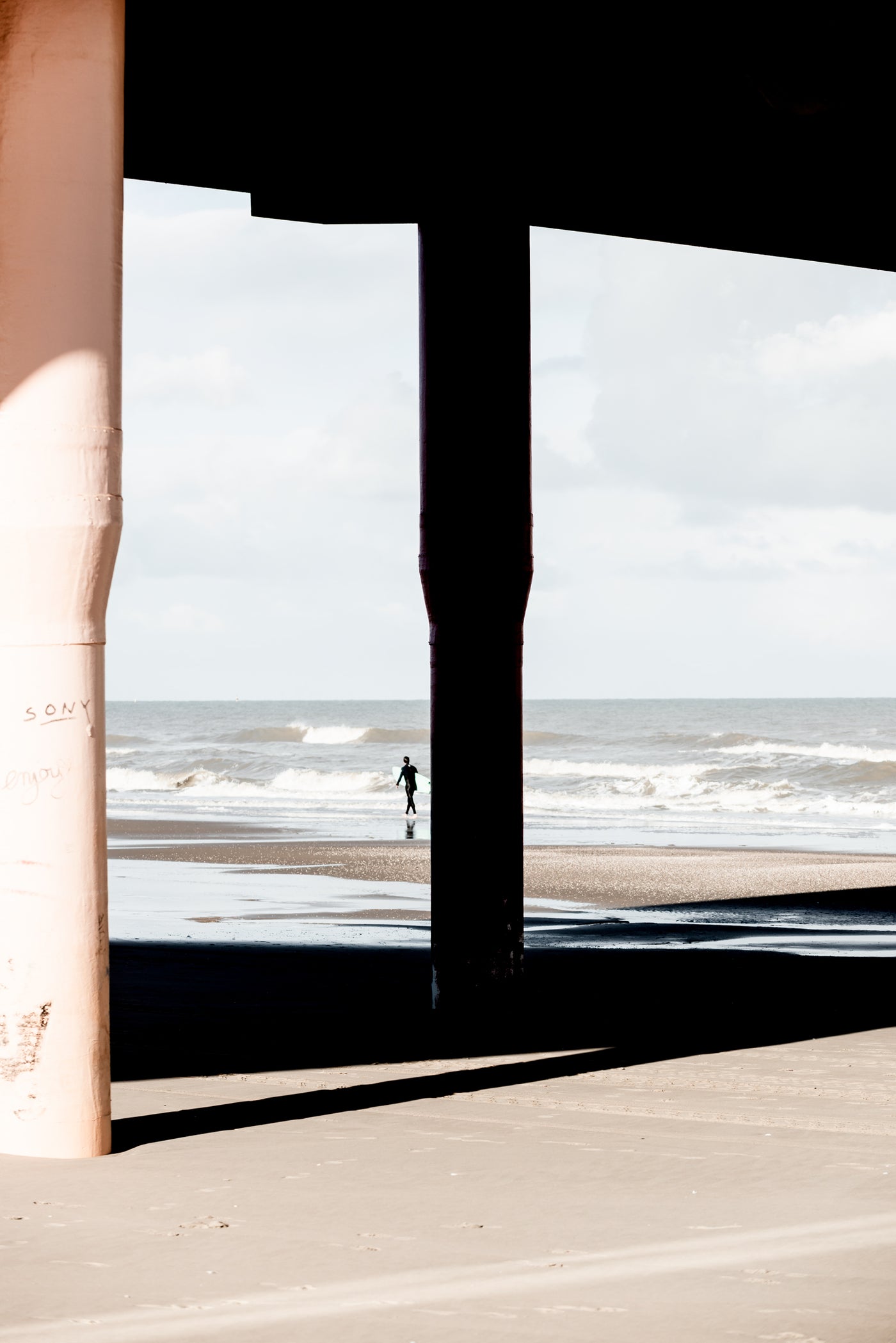 A high-contrast photograph of a beach scene viewed from under a pier, with dark support columns framing the ocean, waves, and a small distant figure on the sand.