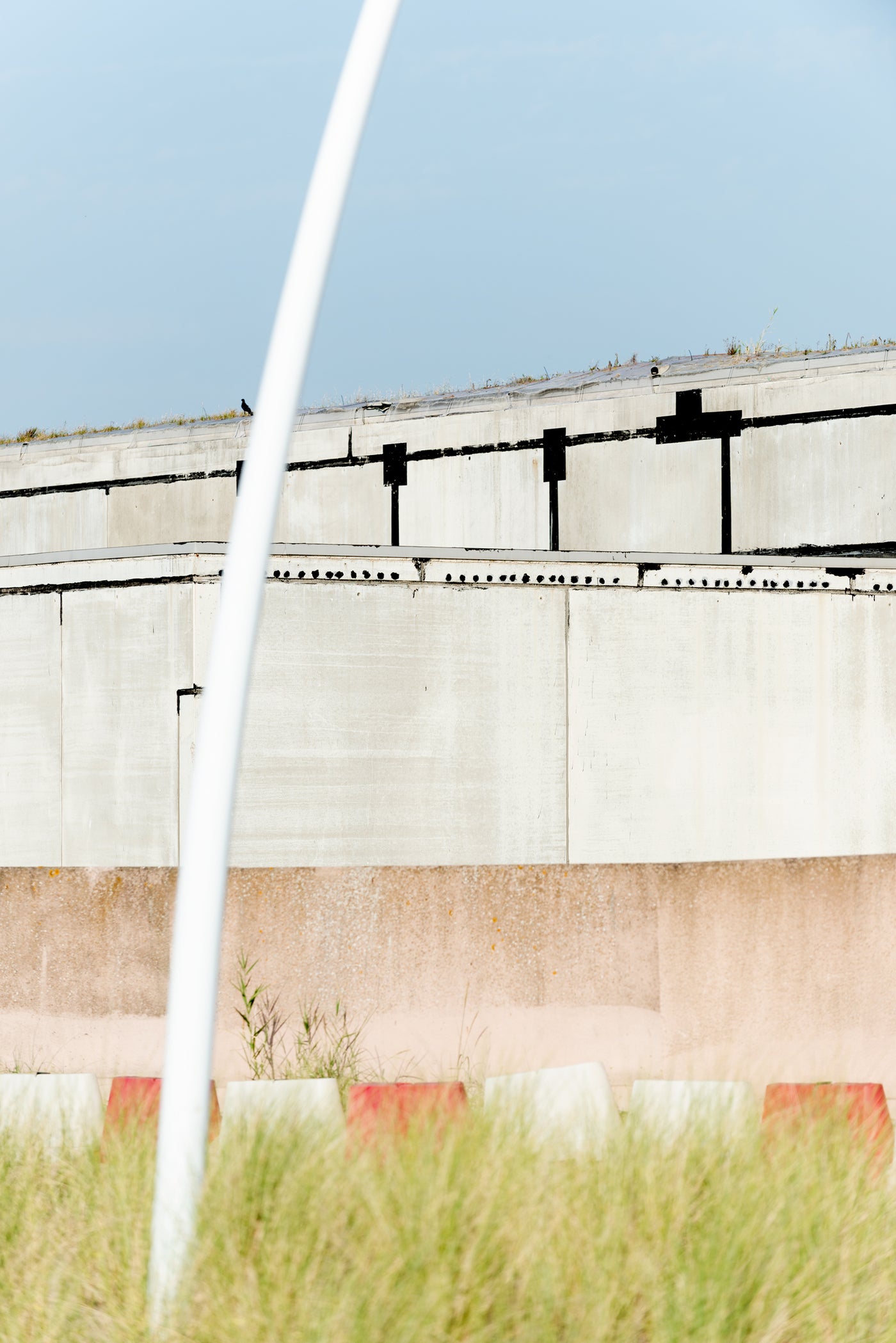 A close-up architectural photograph of a concrete structure with a pale blue sky above and dry grass in the foreground, partially framed by a white vertical pole.