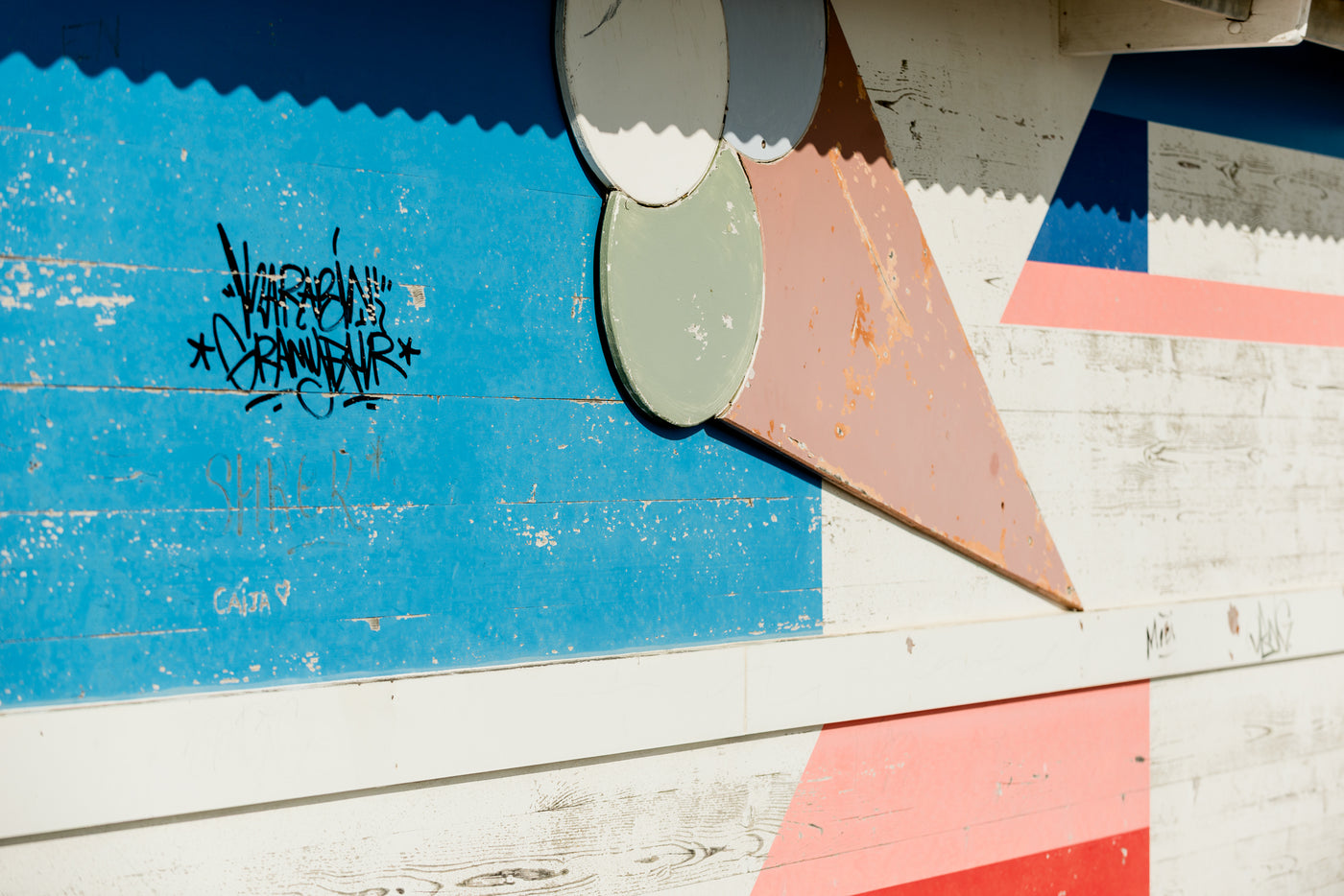 A close-up of a painted wooden wall or door with geometric shapes and graffiti. The visible colors include a large blue panel, white wood planks, a pink triangular shape, a red corner shape, and a gray circular element.