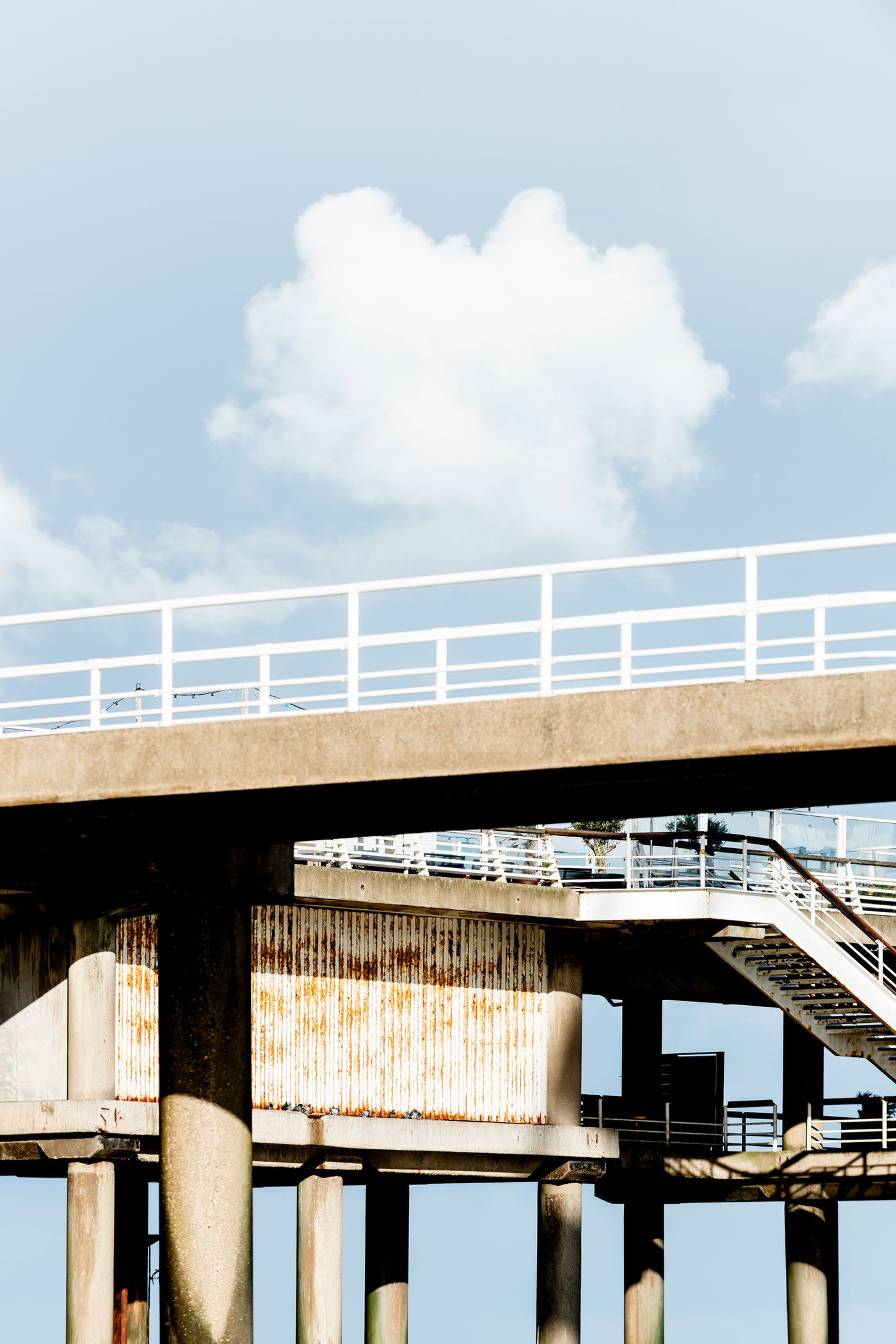 An architectural photograph of a concrete elevated walkway and a staircase at Scheveningen, set against a bright blue sky with a white cloud.