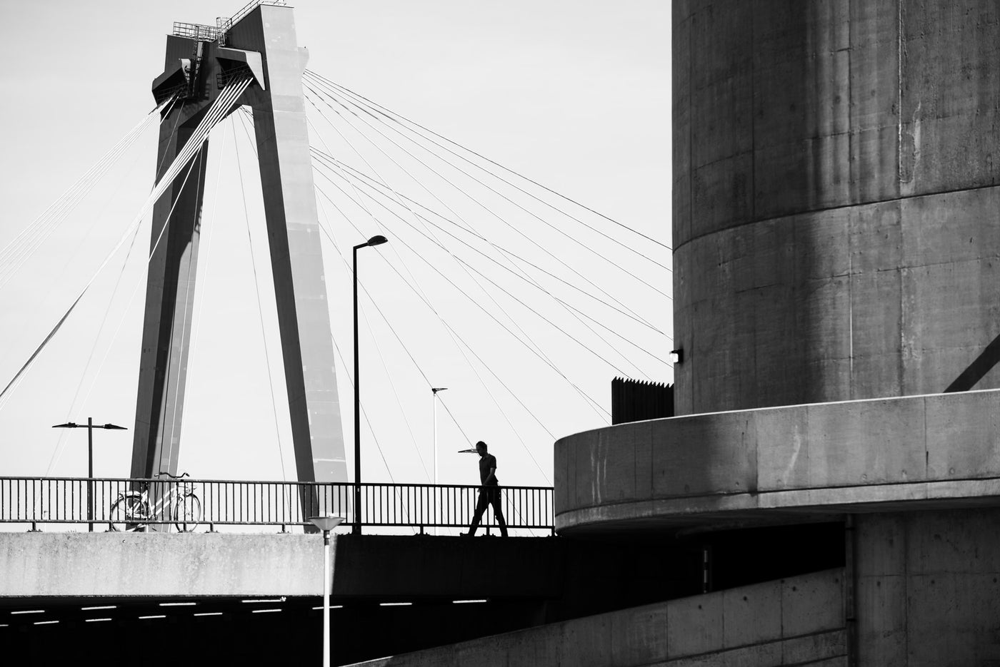 Black and white architectural photograph of a person walking across the Willemsbrug bridge in Rotterdam, framed by large concrete structures.