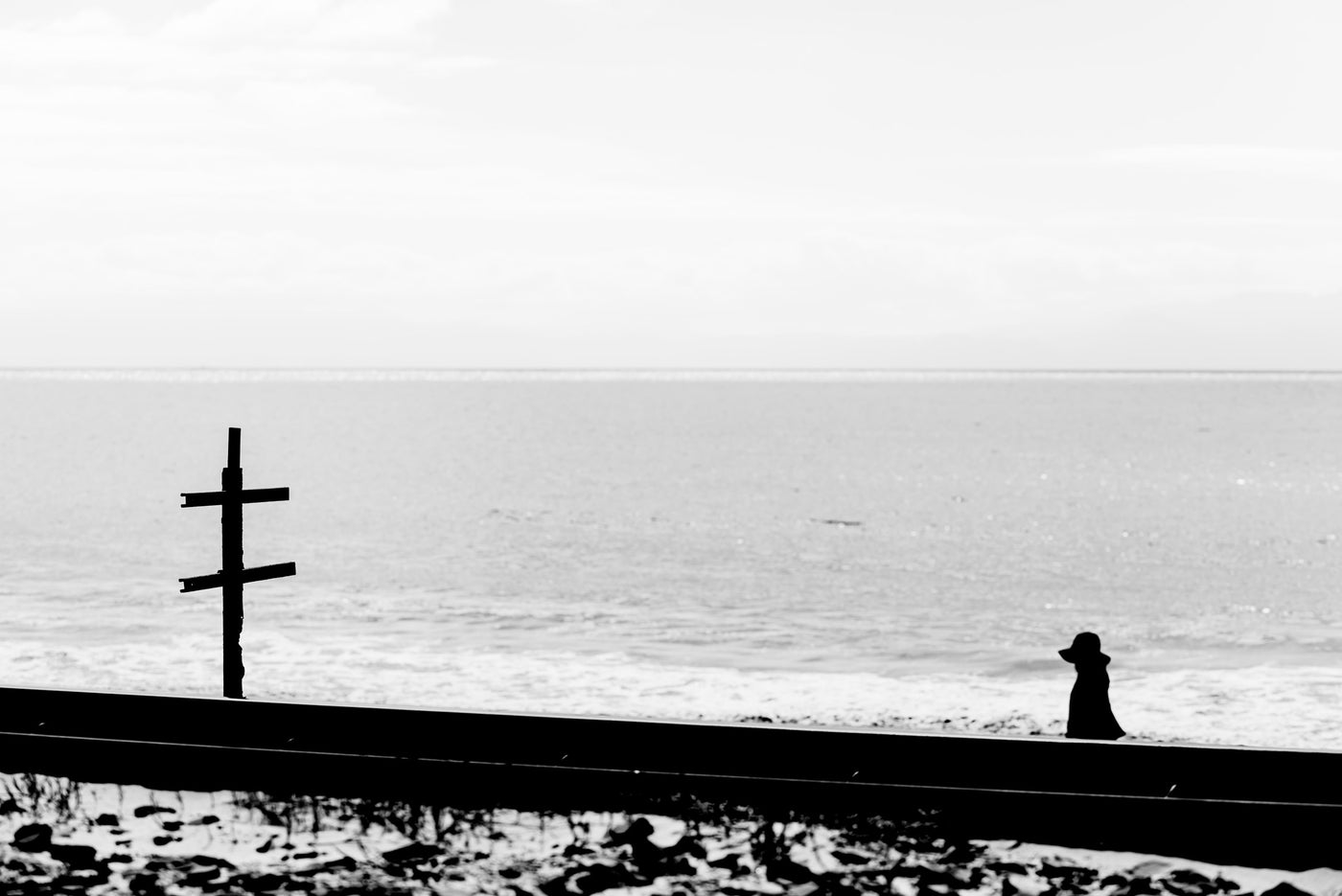 Black and white photograph of a coastal scene with a silhouetted person sitting by the ocean, a wooden post on the left, and a low wall in the foreground.