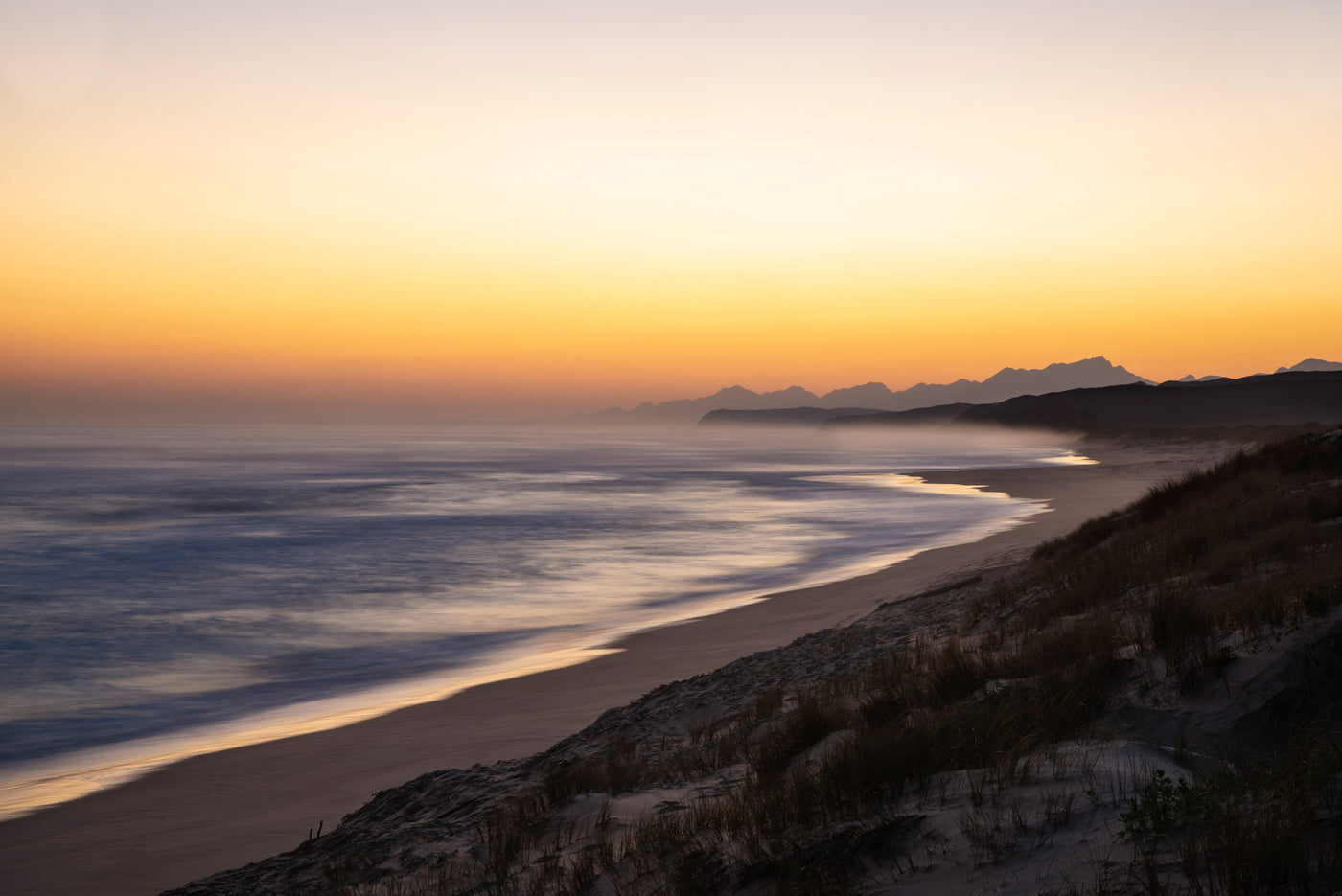 A scenic coastal landscape at sunset with a sandy beach, ocean waves, distant mountains, and a grassy dune in the foreground under a warm orange and yellow sky.