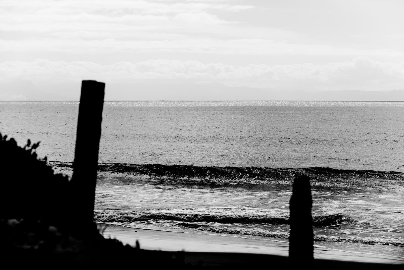 Black and white photograph of an ocean shoreline with waves, a pale cloudy sky, and dark silhouetted posts in the foreground.