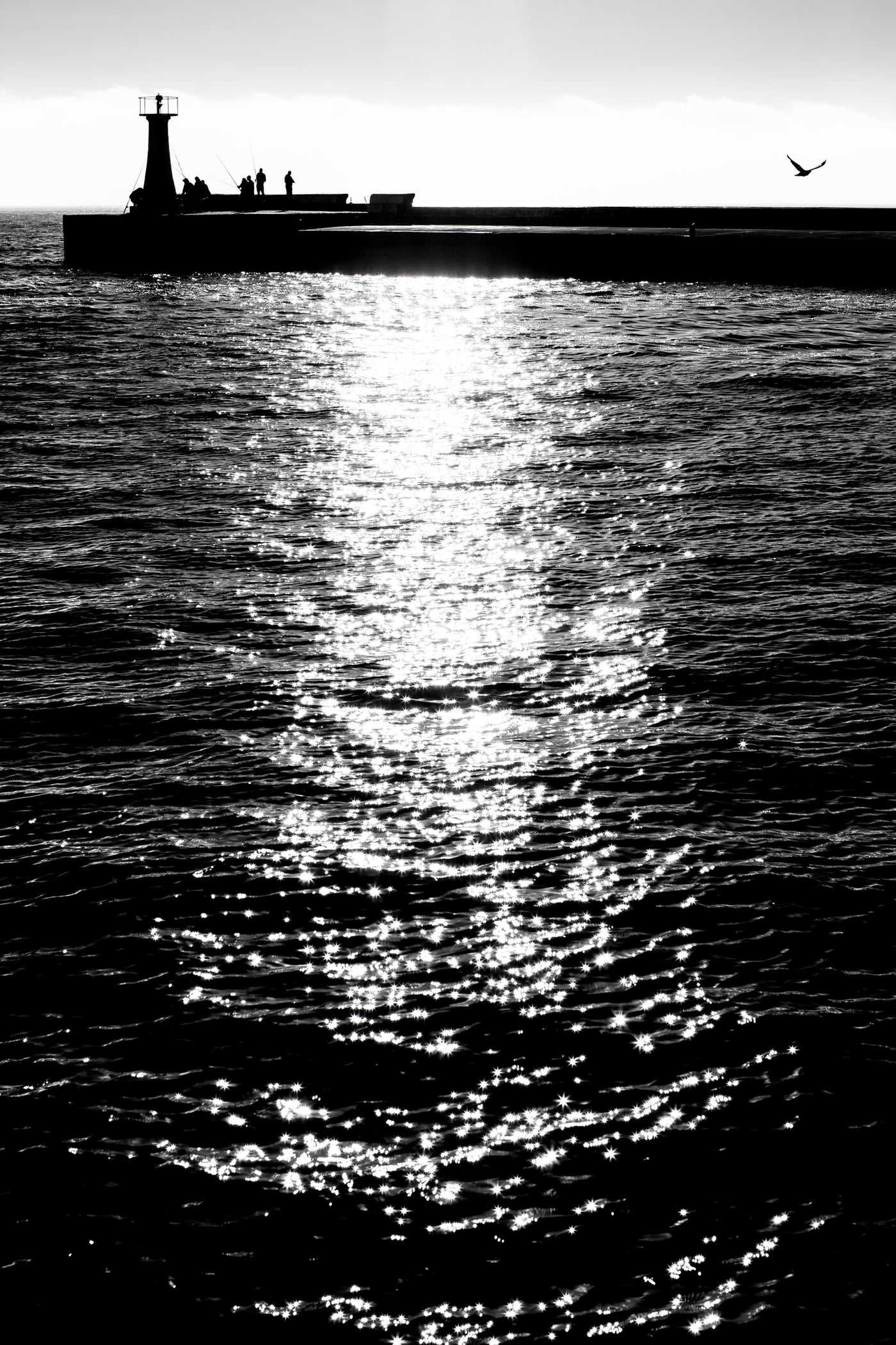 Black and white photograph the pier at Kalk bay extending into the water, with a lighthouse silhouette on the left, a bird in the sky, and bright sunlight reflecting across the water surface.