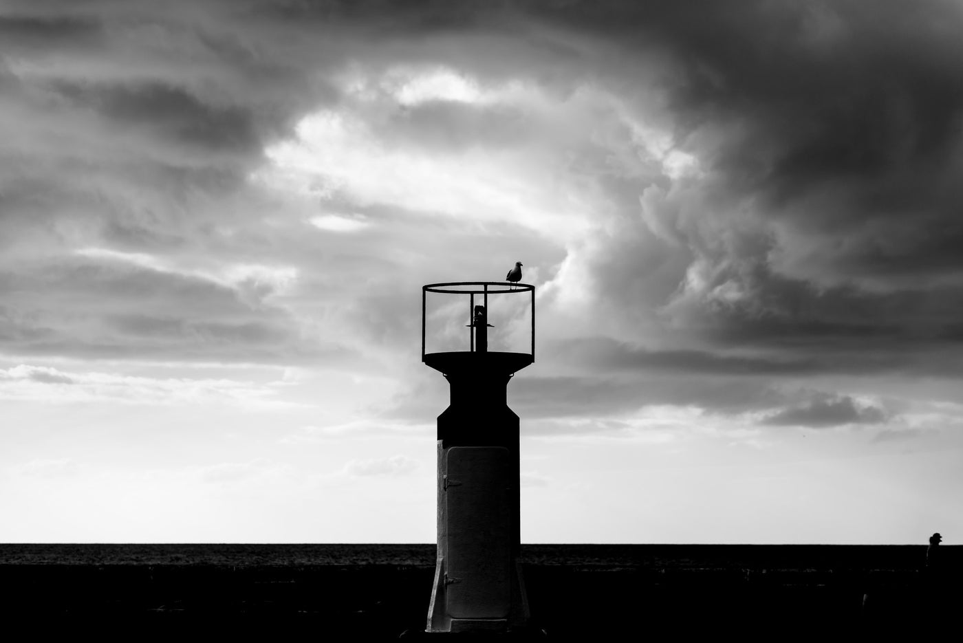 Black and white landscape photograph of the lighthouse at Kalk Bay silhouette against a dramatic cloudy sky, with birds perched on top.