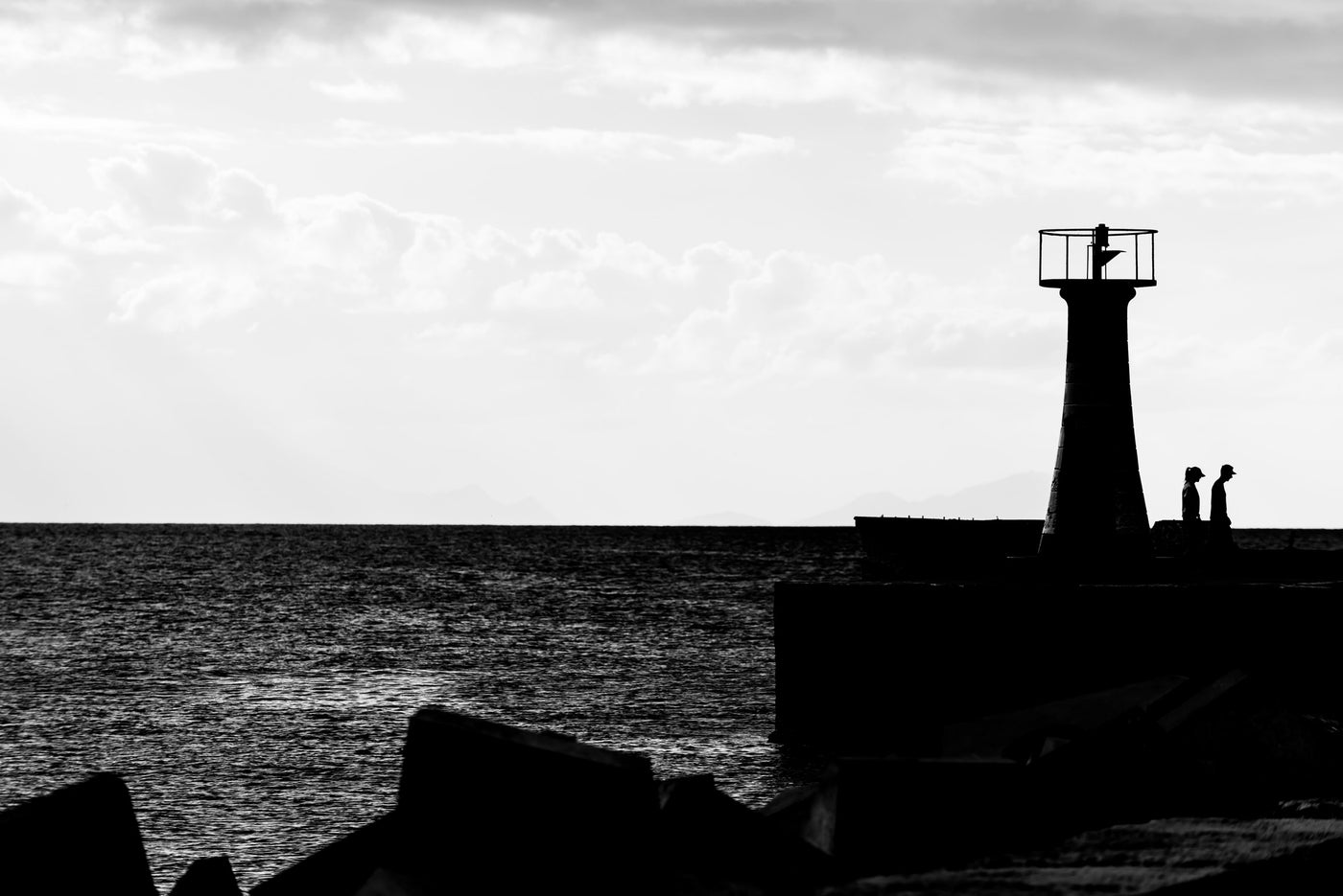 Black and white photograph of Kalk bay lighthouse silhouette on a breakwater with ocean water and cloudy sky, with small human figures visible near the lighthouse.