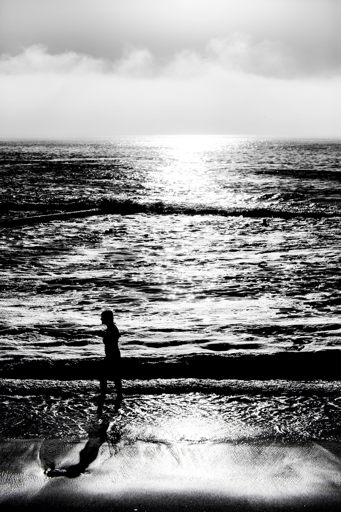 Black and white photograph of a person standing at St James tidal pool at the shoreline, with waves, a bright sun reflection on the water, and a cloudy sky.