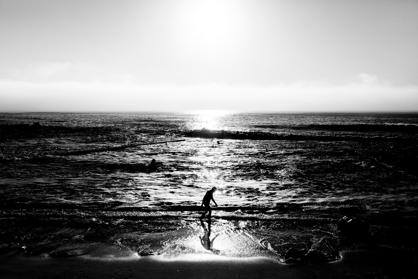 Black and white photograph of a person walking at St James Tidal Pool at sunset, with the sun low over the horizon and bright reflections on the water.