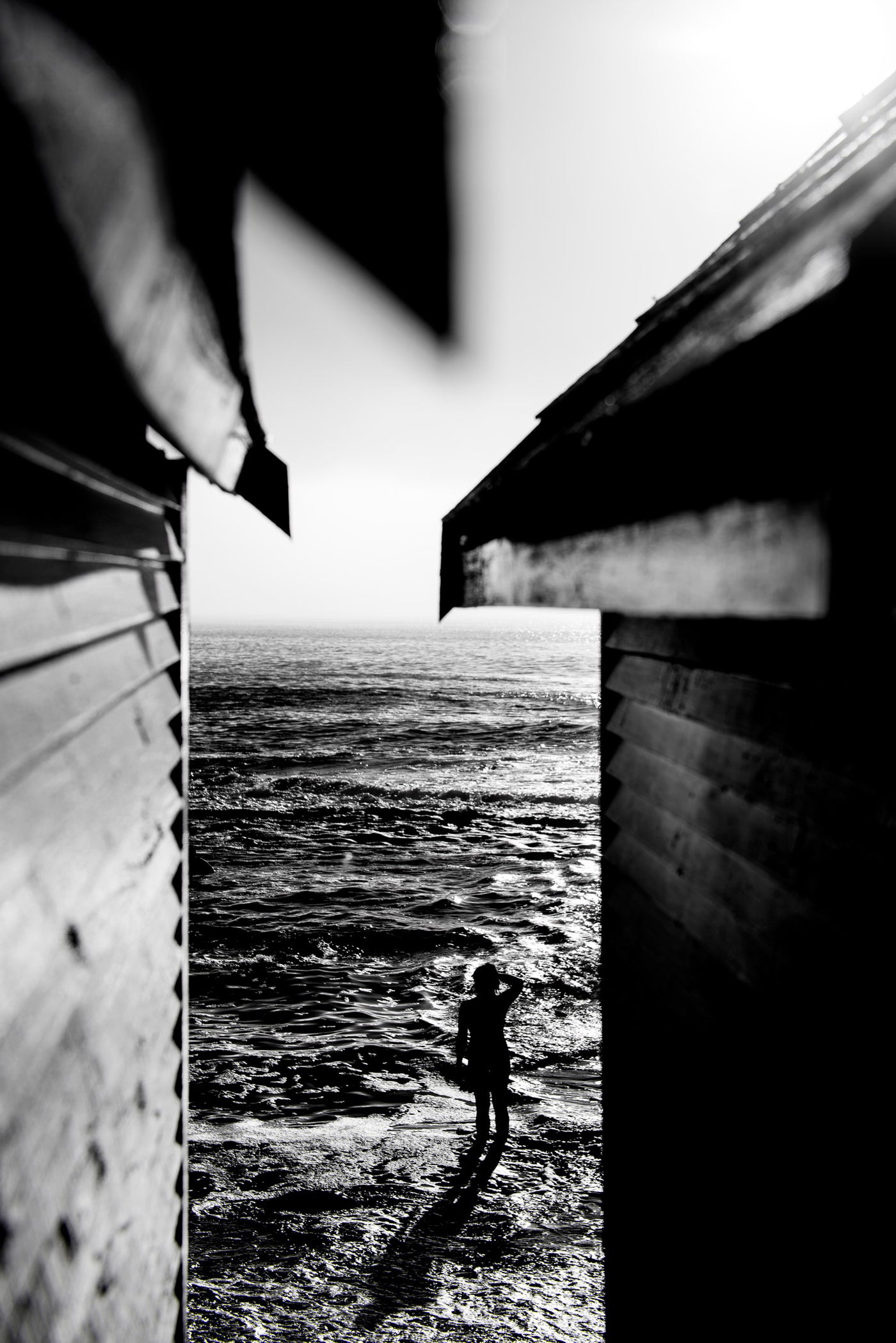 Black and white photograph of a person standing in shallow water between two changing huts at St James, Cape Town, with the ocean visible in the background.