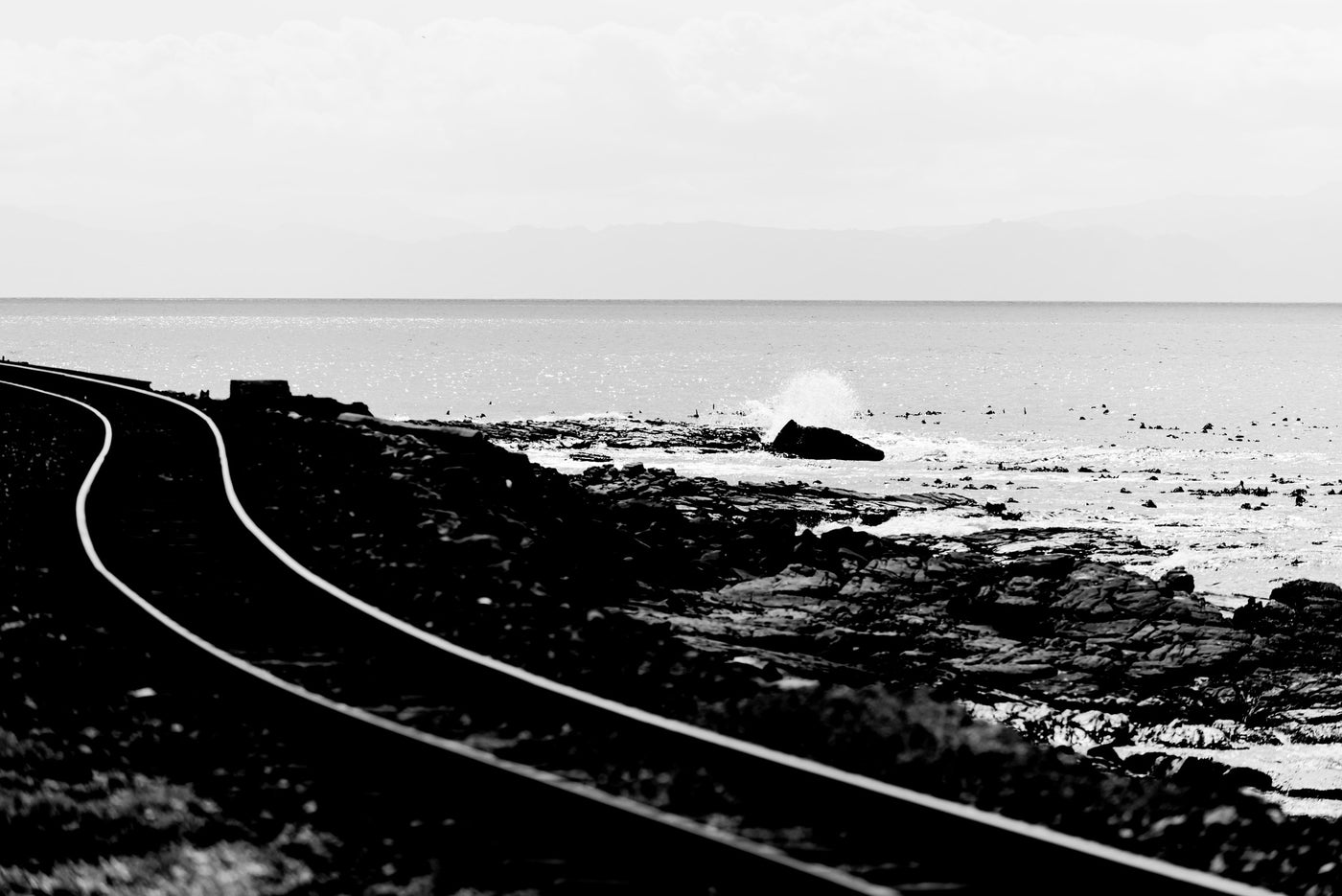 Black and white landscape photograph of a coastal scene with a curved railway track running along the rocky deep south shoreline toward Simonstown.