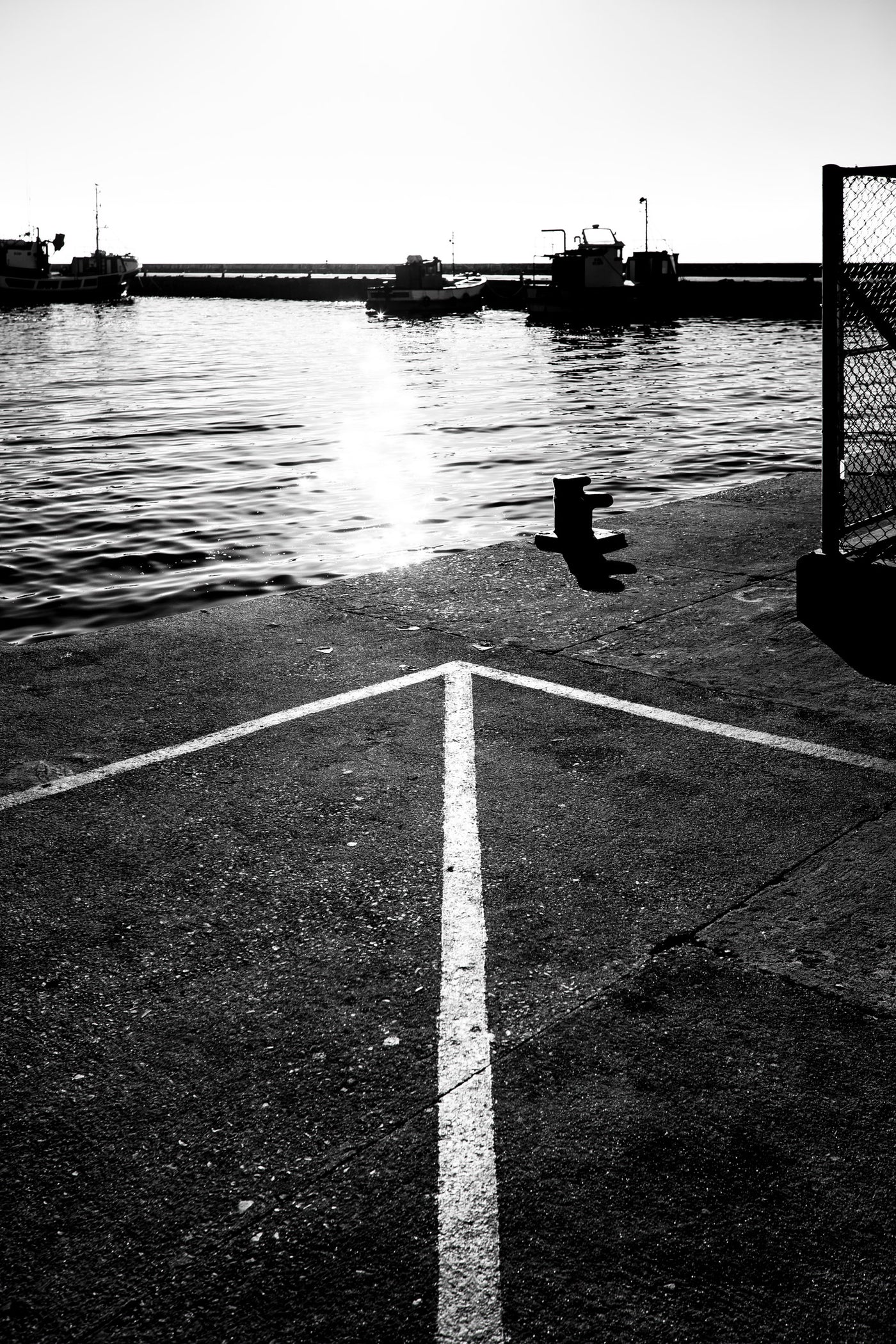 Black and white photograph of a harbor scene at Kalk Bay with water, boats, a dock, a chain-link fence, and a painted white directional line on the pavement.